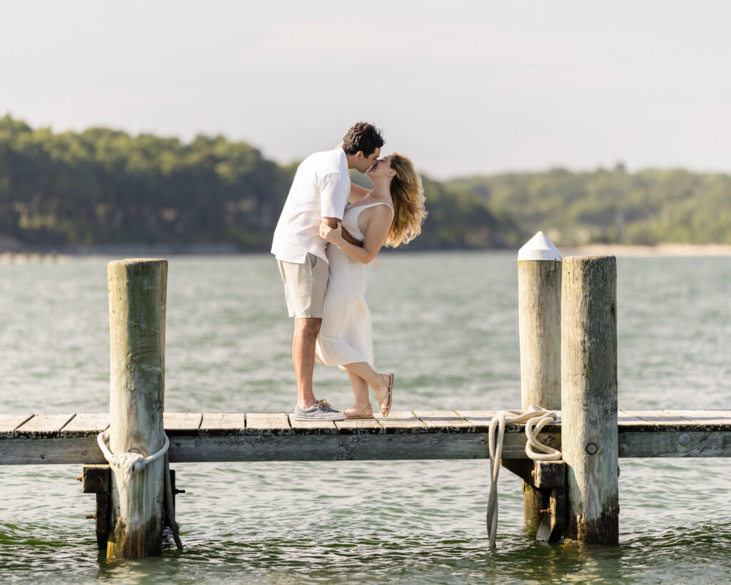 James lifts Kristen into a kiss as they stand barefoot on the dock, water behind them.