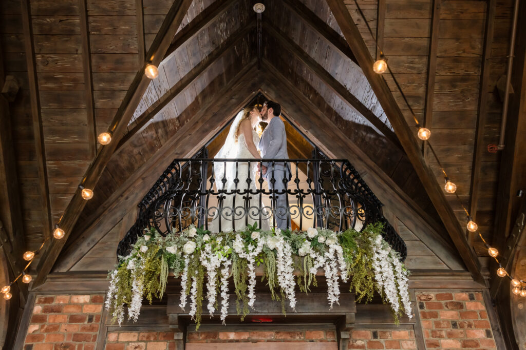 Bride standing on the grand balcony of Riviera Waterfront Mansion, framed by floral arrangements and rustic architecture.