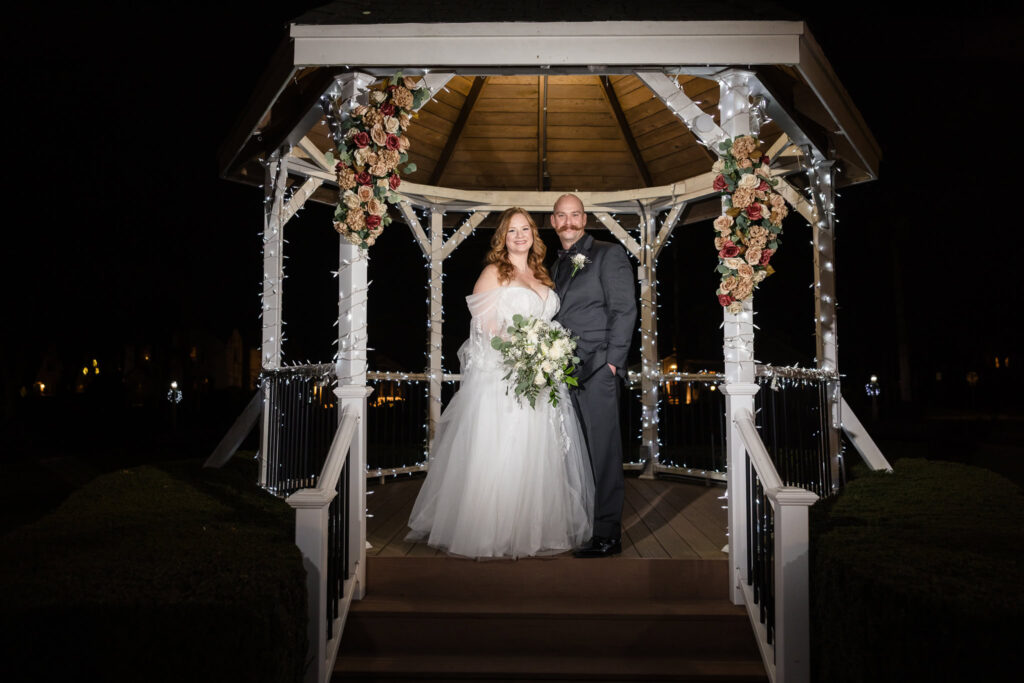 Night portrait of bride holding bouquet beneath Stewart Manor’s white gazebo, glowing with romance.