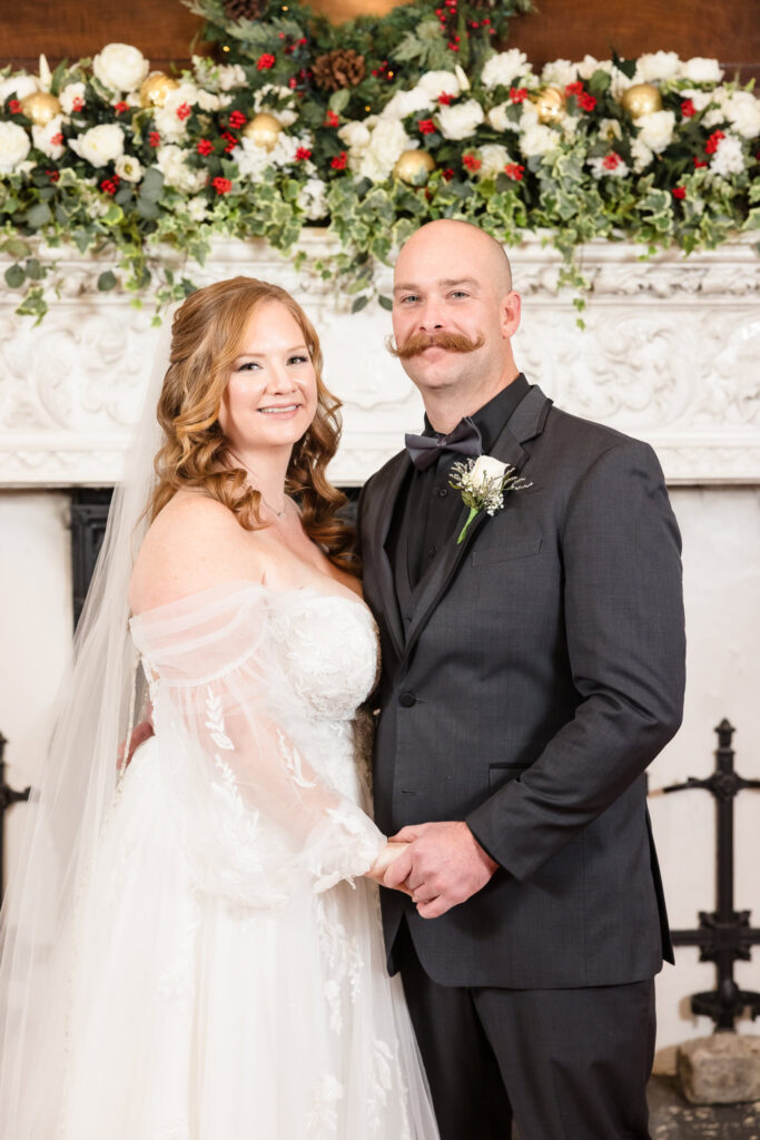 Bride and groom smiling together in front of Stewart Manor’s historic fireplace, surrounded by florals.