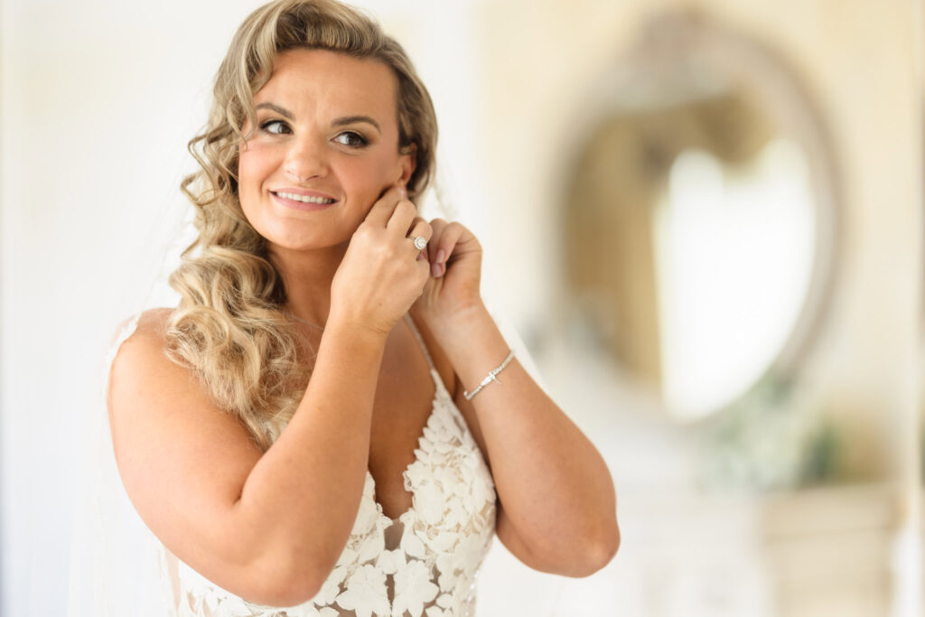 Bride smiling as she puts on her earrings, framed by soft natural light and elegant bridal details at Stonebridge Country Club in Smithtown, NY.