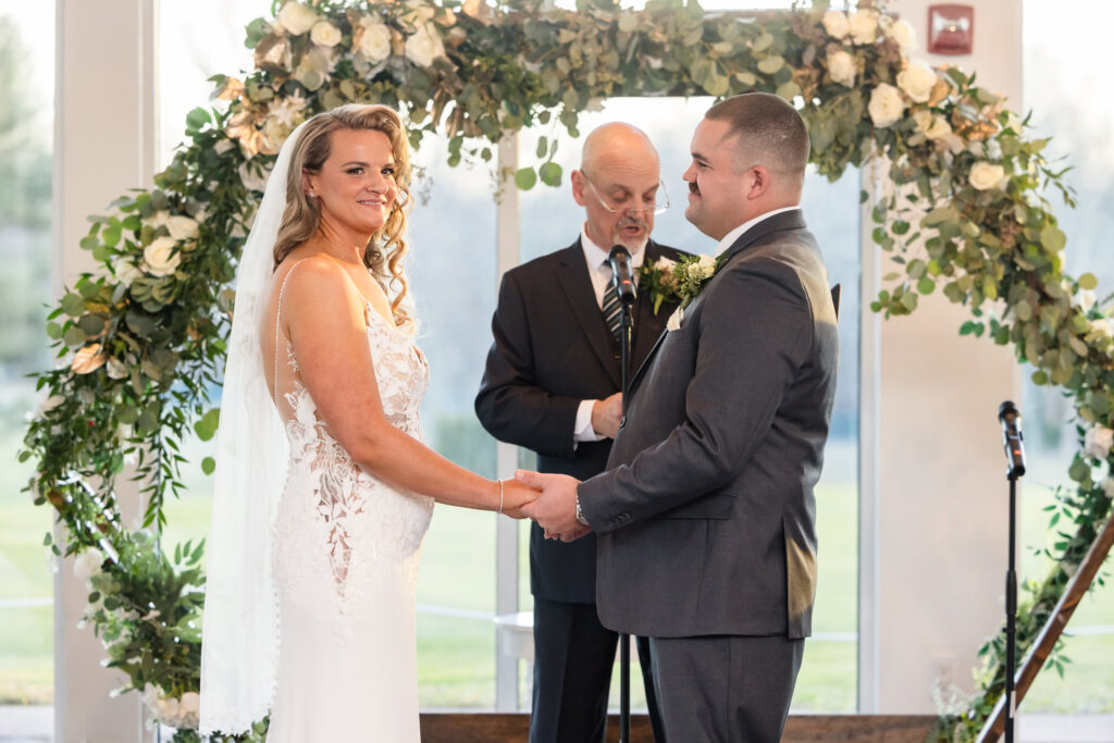 Bride and groom exchange vows under a lush floral arch during their indoor ceremony at Stonebridge Country Club, surrounded by soft natural light and emotional presence.

