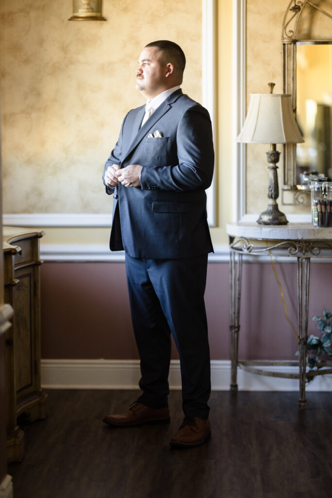 Groom stands in quiet reflection before the ceremony, lit by warm window light in the groom’s suite at Stonebridge Country Club.