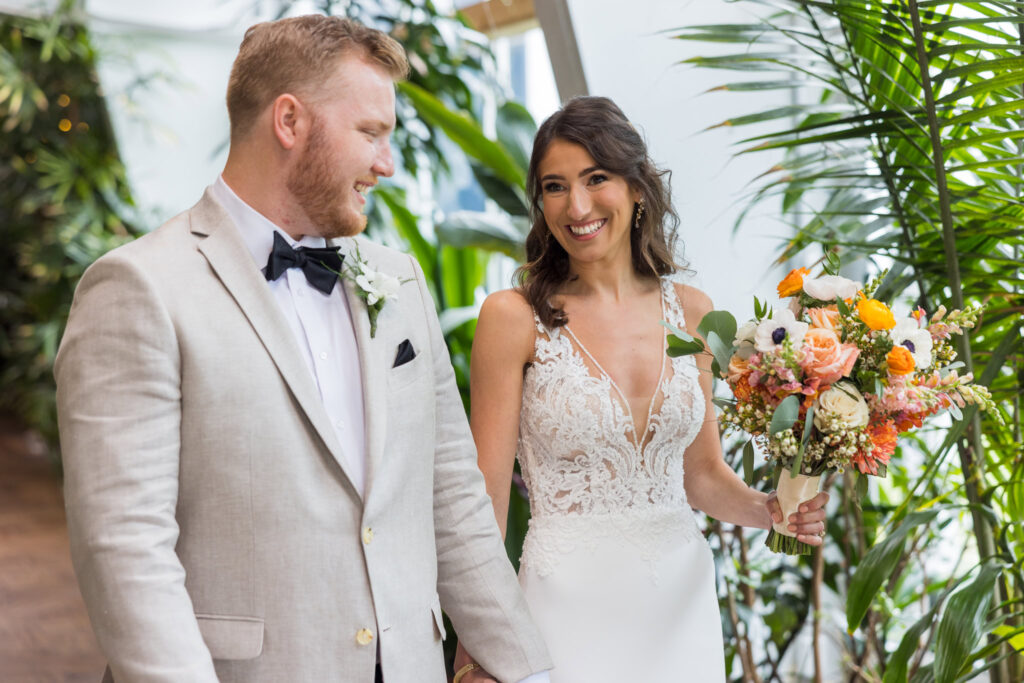 The bride and groom walk hand-in-hand through Flowerfield’s greenhouse, framed by lush tropical foliage and the soft joy of just-said vows.

