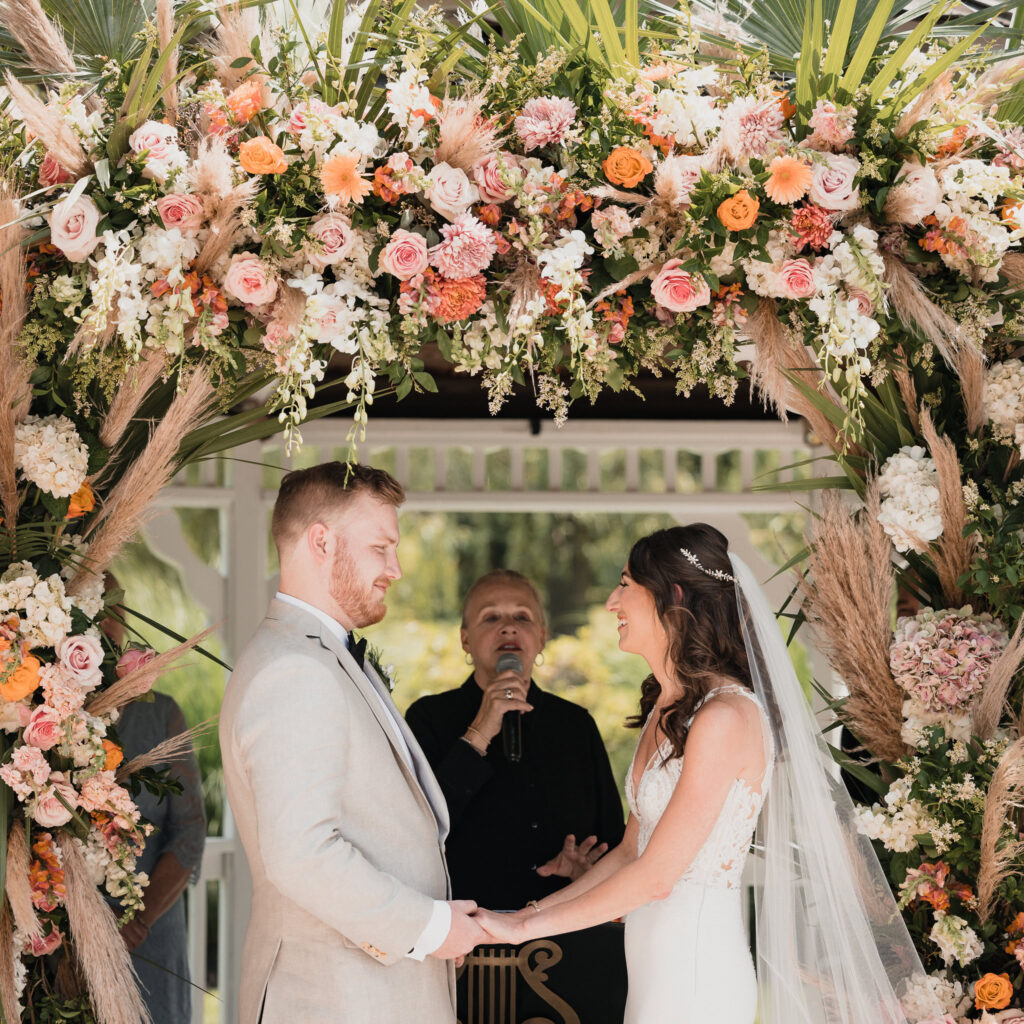 Bride and groom hold hands beneath a lush floral arbor during their outdoor ceremony at Flowerfield, surrounded by vibrant blooms and sunlight as they exchange heartfelt vows.

