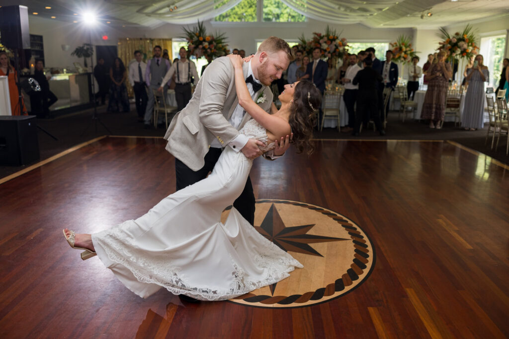 The groom dips his bride during their first dance as guests cheer around them, the rich wood floors and floral-filled ballroom setting the stage for a perfect celebration.

