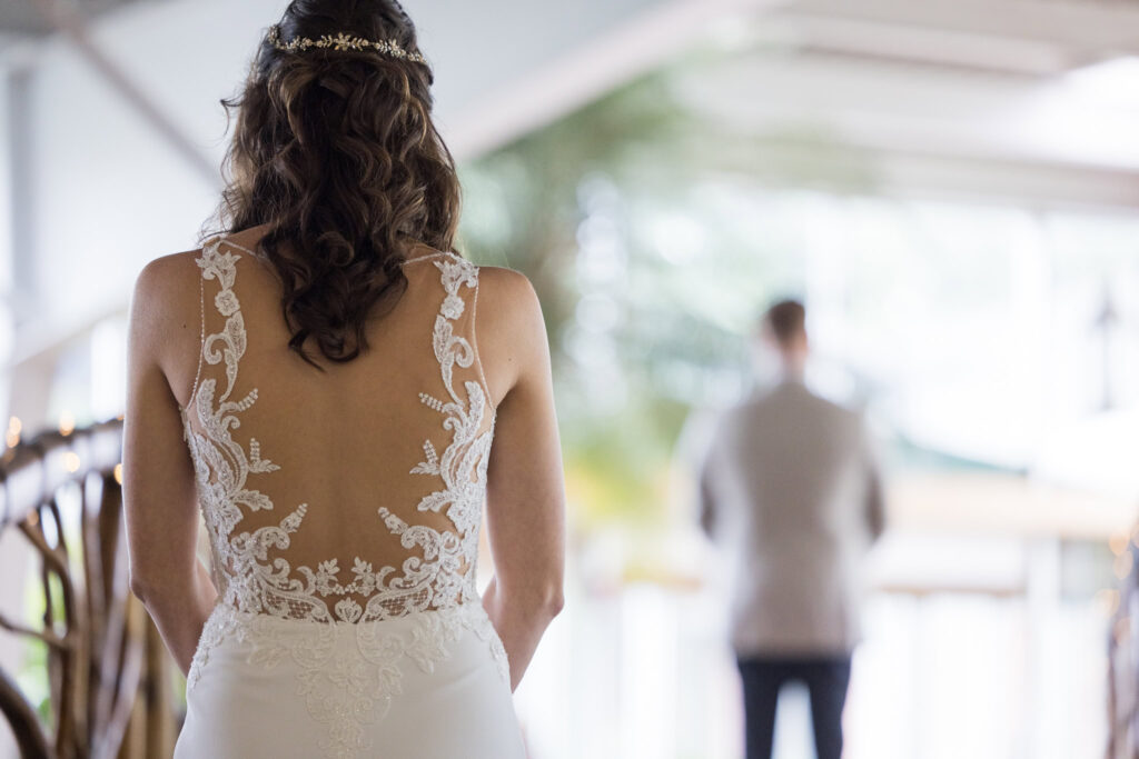 The bride approaches her groom from behind inside Flowerfield’s greenhouse, her lace gown glowing softly as she prepares for their emotional first look.

