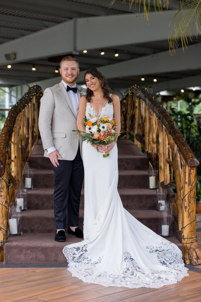 The bride and groom pose together on the candlelit wooden staircase inside Flowerfield’s greenhouse, her lace train cascading down the steps in timeless elegance.

