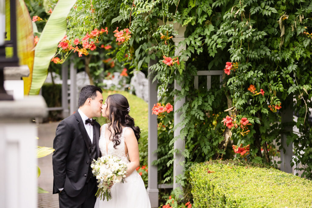 Bride and groom share a romantic kiss beneath a lush floral arbor at Watermill Caterers in Smithtown, NY, surrounded by vibrant greenery and cascading orange blossoms.