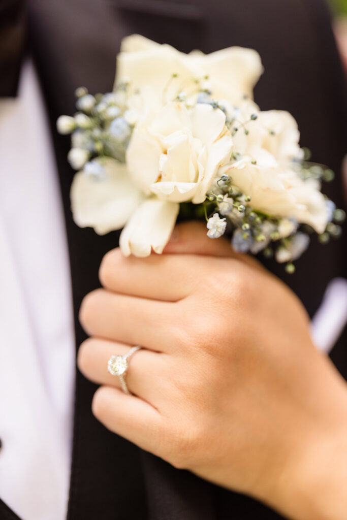 Close-up of bride’s hand with diamond engagement ring gently resting on groom’s chest, highlighting an elegant white boutonnière against a classic black tuxedo.