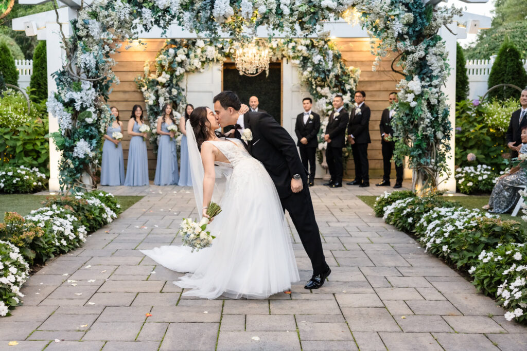 Groom dips and kisses bride during their wedding ceremony at Watermill Caterers, surrounded by white floral arches, lush garden blooms, and their bridal party in soft blue and black attire.