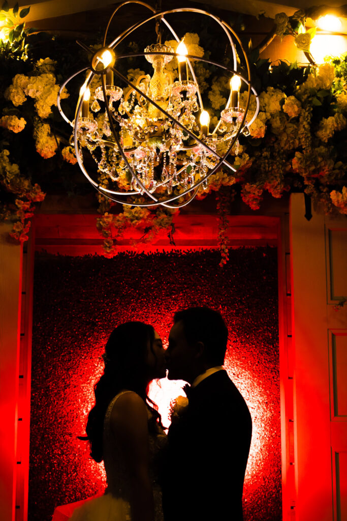 Bride and groom share a romantic kiss in silhouette beneath a glowing chandelier at Watermill Caterers, framed by dramatic red lighting and lush floral decor.