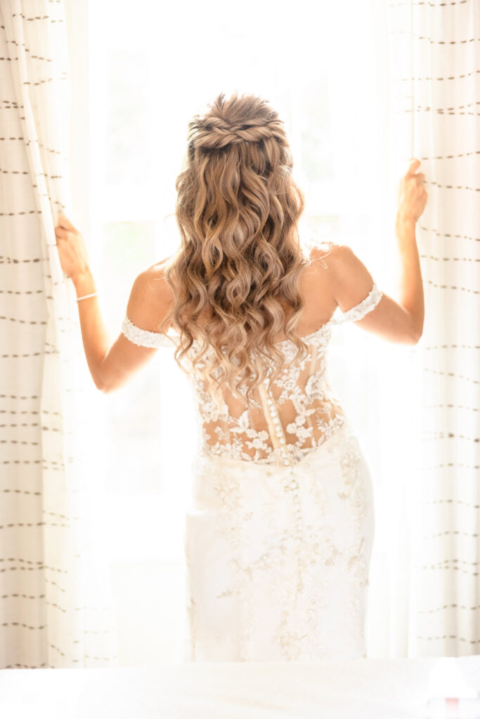 Bride gazing out of a sunlit window, back of lace gown in focus