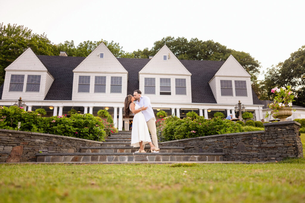 Christine and Matt standing together in front of the Centerport Yacht Club, framed by manicured landscaping.