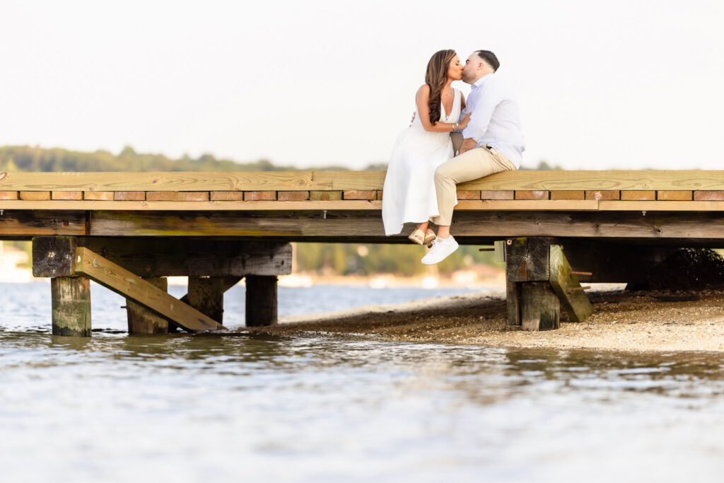 Christine and Matt sitting at the edge of a wooden dock, feet dangling over the water as they look out at the harbor.
