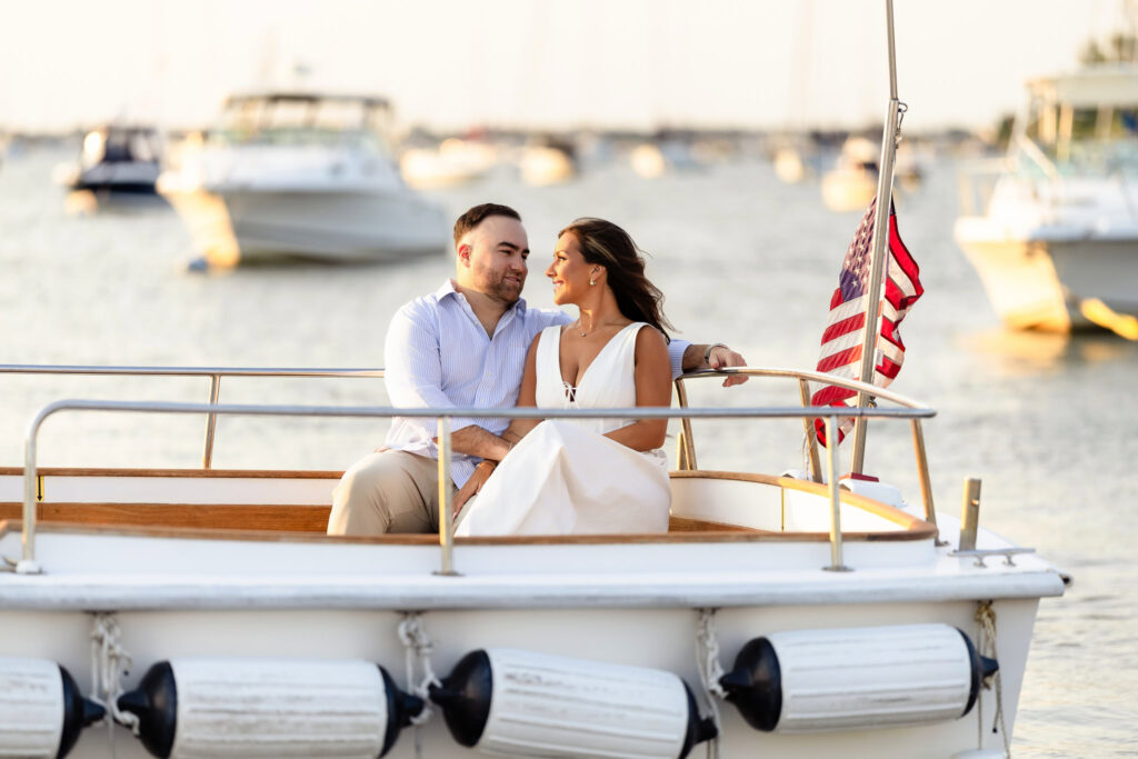 Christine and Matt sitting together at the bow of a boat, sailboats and harbor reflections behind them.
