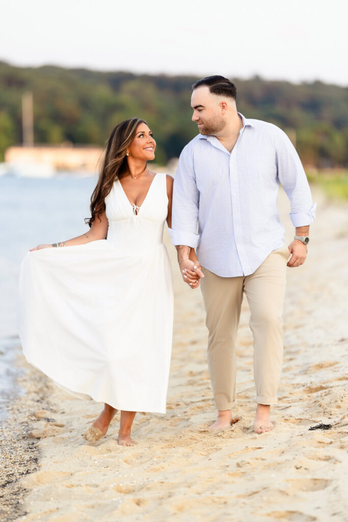 Christine and Matt walking barefoot along the sandy shoreline, holding hands with soft waves at their feet.