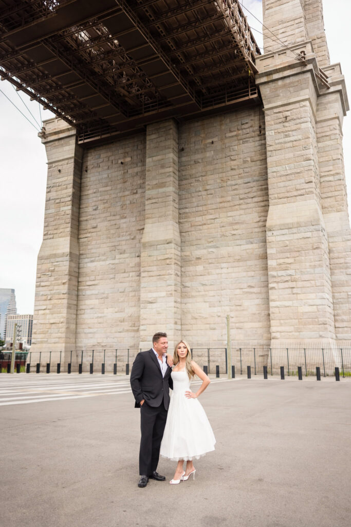 Couple standing beneath the stone archway of the Brooklyn Bridge during their DUMBO engagement session.