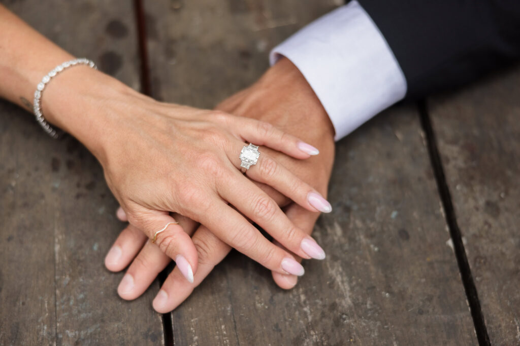 Close-up of couple’s hands with a radiant emerald-cut engagement ring on a rustic wooden table.
