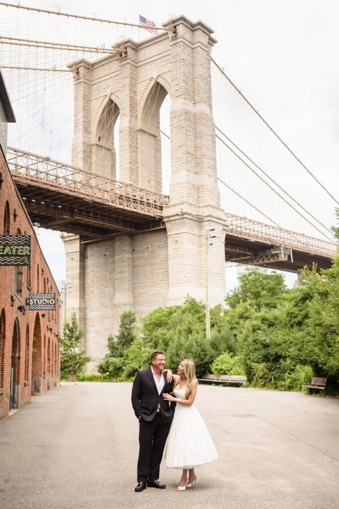 Couple standing on a quiet path in DUMBO with the Brooklyn Bridge rising in the background.