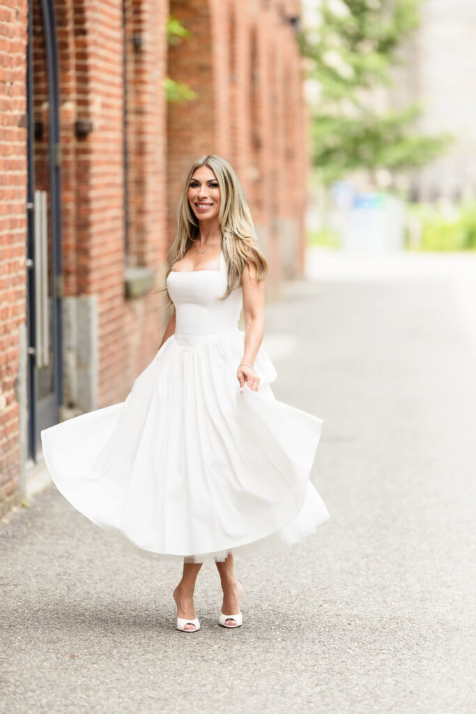 Woman in a white dress twirling joyfully near a red brick building in DUMBO, Brooklyn.