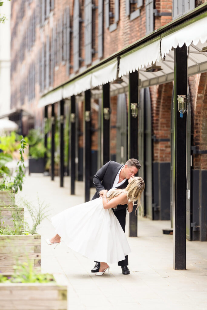 Man dipping his fiancée for a kiss along a shaded walkway in DUMBO with red brick architecture behind them.