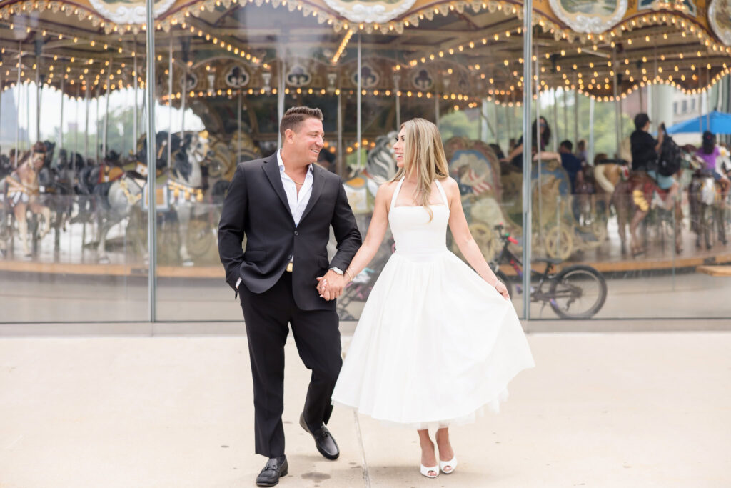 Couple holding hands and smiling in front of Jane’s Carousel during their DUMBO engagement session.