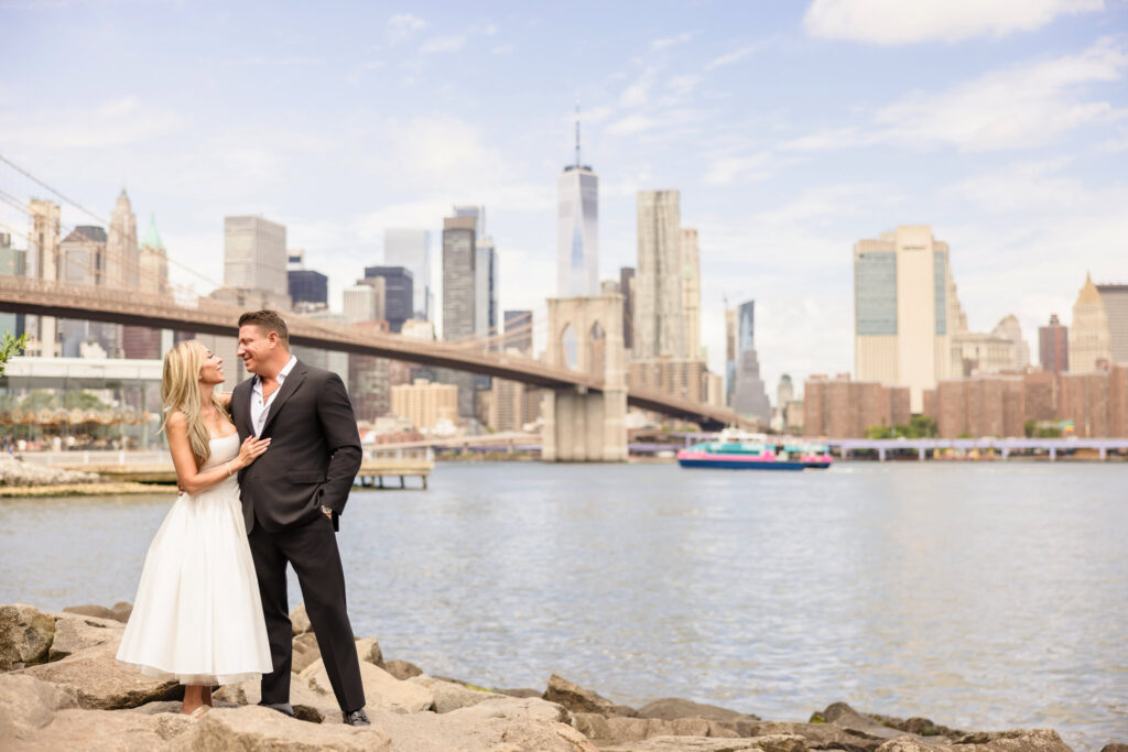 Engaged couple standing on rocks at Pebble Beach with the Brooklyn Bridge and Manhattan skyline behind them.