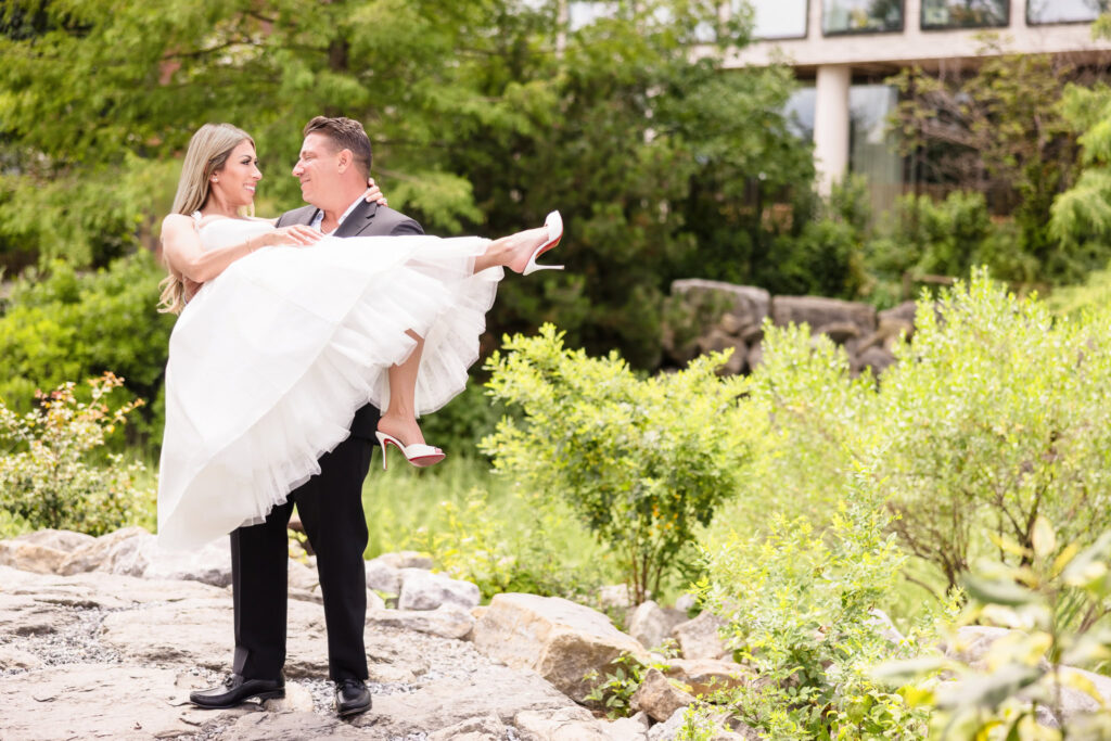 Man lifting his fiancée in a white dress while standing on rocks surrounded by greenery in Brooklyn Bridge Park.