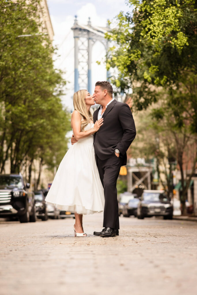 Couple kissing on a cobblestone street in DUMBO with the Manhattan Bridge rising in the background.