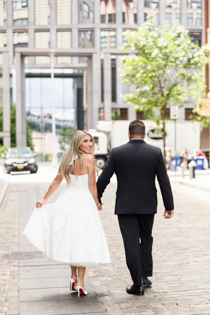 Woman smiling over her shoulder while walking hand-in-hand with her fiancé on a cobblestone street in DUMBO.