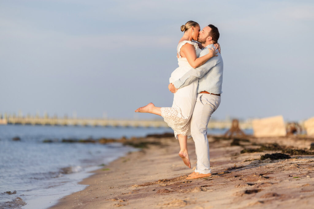 Couple kissing on the sandy shore at Fire Island, with the man lifting the woman in the air near the water.