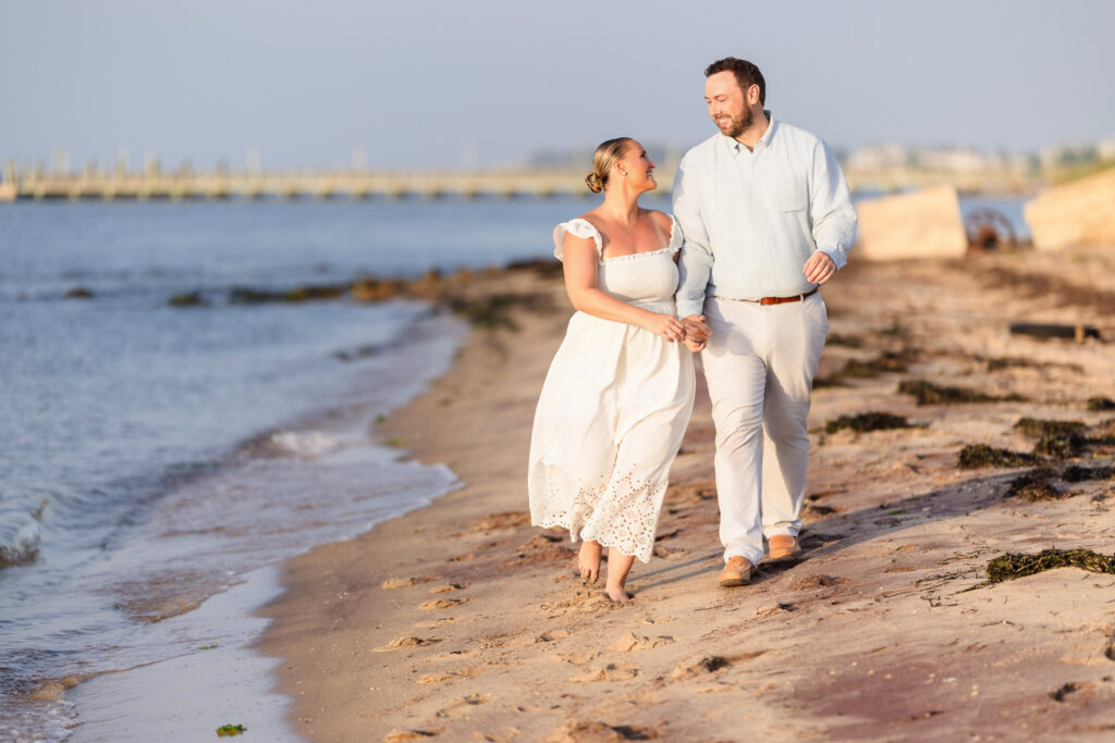 Couple walking barefoot along the beach at Fire Island, holding hands and smiling at each other.