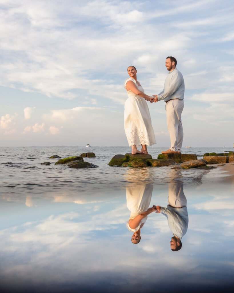 Couple holding hands on rocks by the ocean at Fire Island, with their reflection visible in the water.