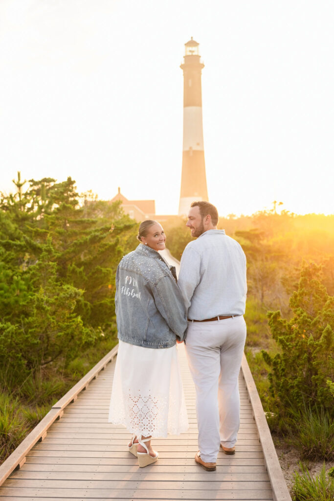 Couple holding hands on the boardwalk at Fire Island Lighthouse during sunset, bride wearing custom denim jacket.
