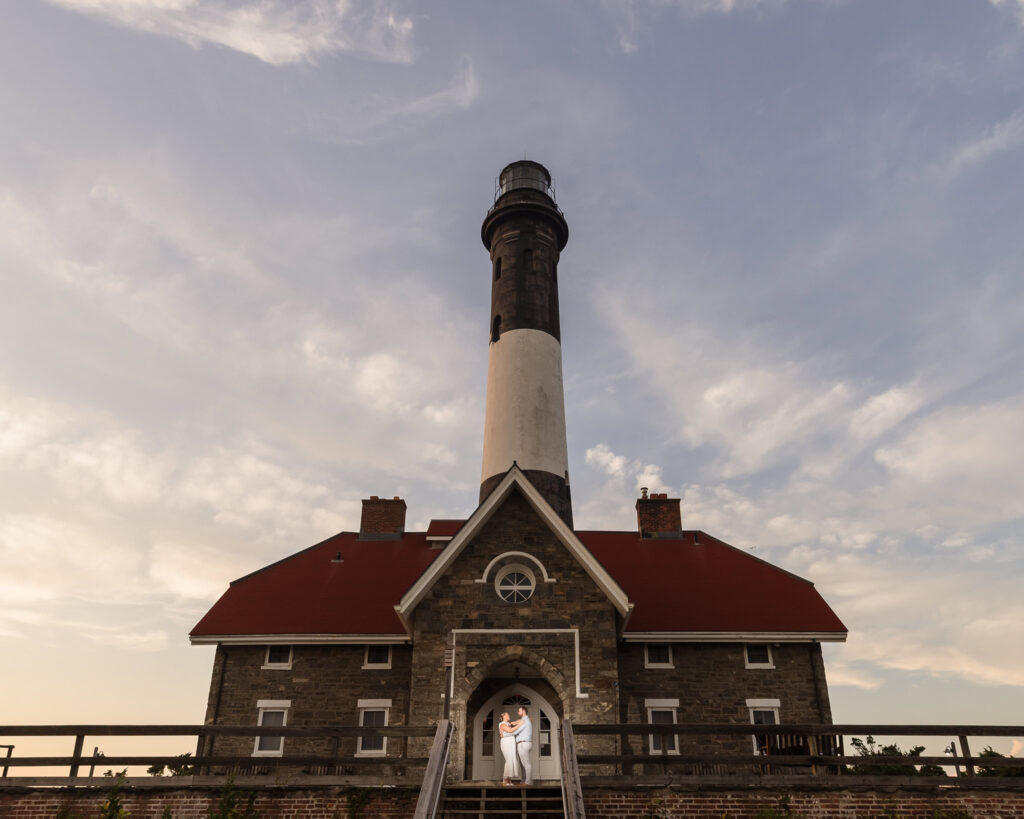 Couple embracing at the entrance of the Fire Island Lighthouse, framed by the historic stone building at sunset.