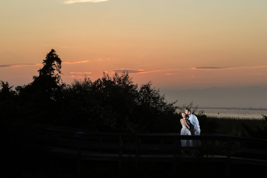 Couple standing on a boardwalk at Fire Island during twilight, with the sky glowing in shades of orange and pink.