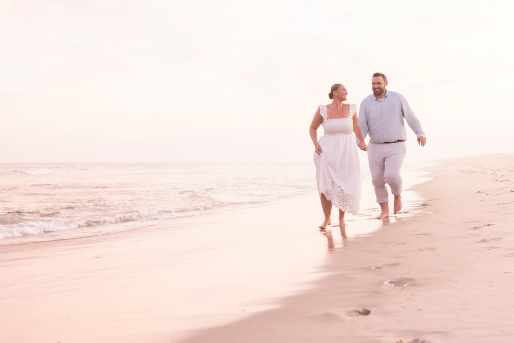 Couple walking hand in hand along the shoreline at Fire Island with soft pastel light reflecting on the sand.