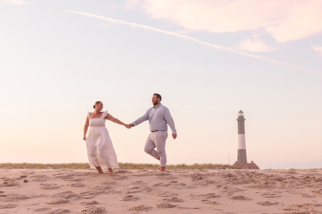 Couple walking hand in hand on the sand at Fire Island with the lighthouse in the background at sunset.