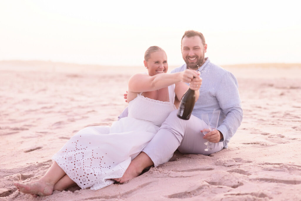 Couple sitting on the sand at Fire Island, laughing while opening a champagne bottle during their engagement session.