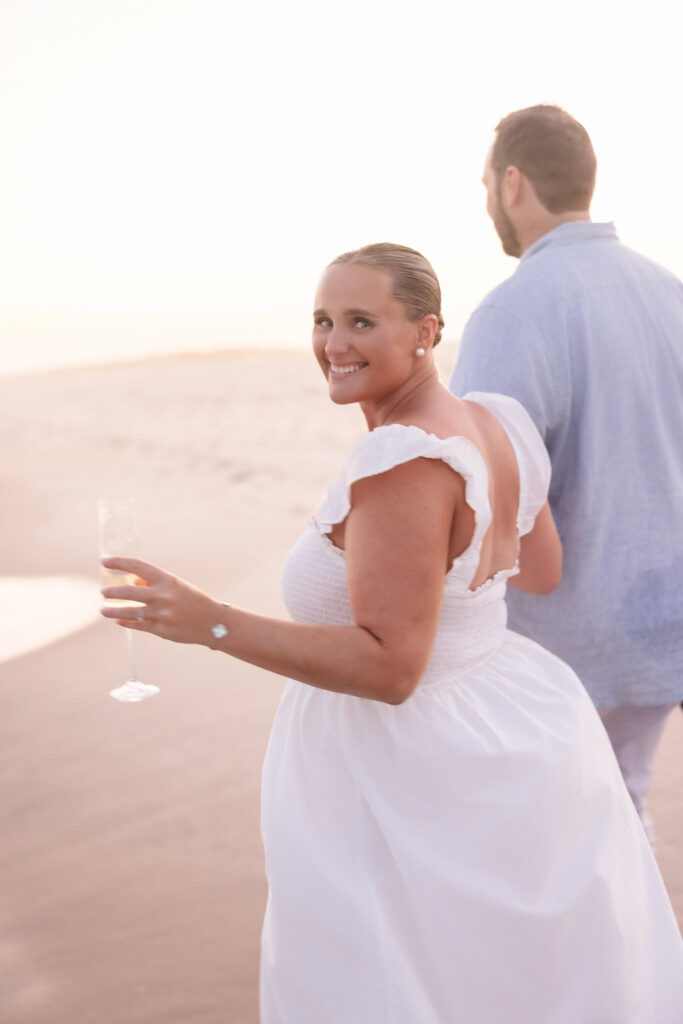 Woman smiling at the camera while holding a champagne glass, walking on the beach with her partner.