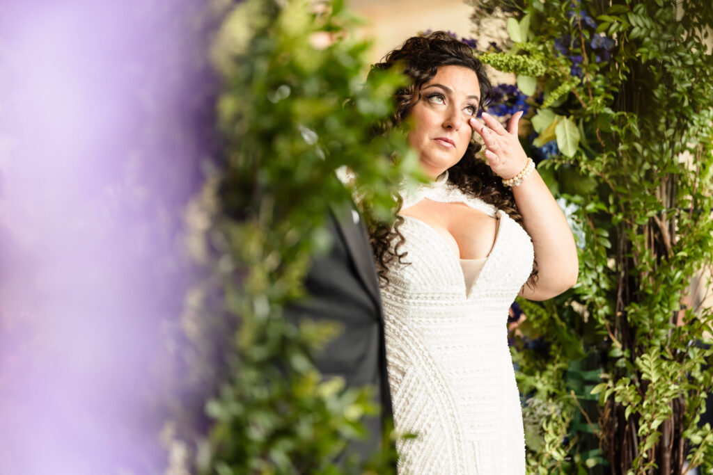 Bride standing alone in front of a floral arch, smiling gently with one hand in her hair as purple florals frame her.
