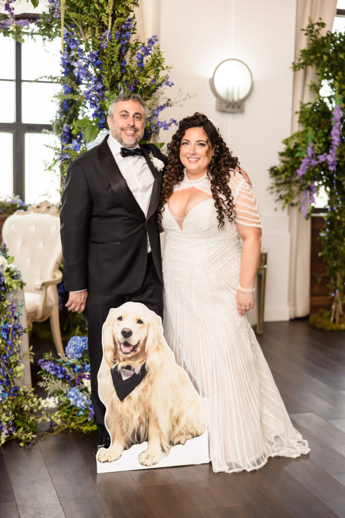 Bride and groom posing with their dog’s cardboard cutout beneath the floral chuppah, smiling joyfully together.