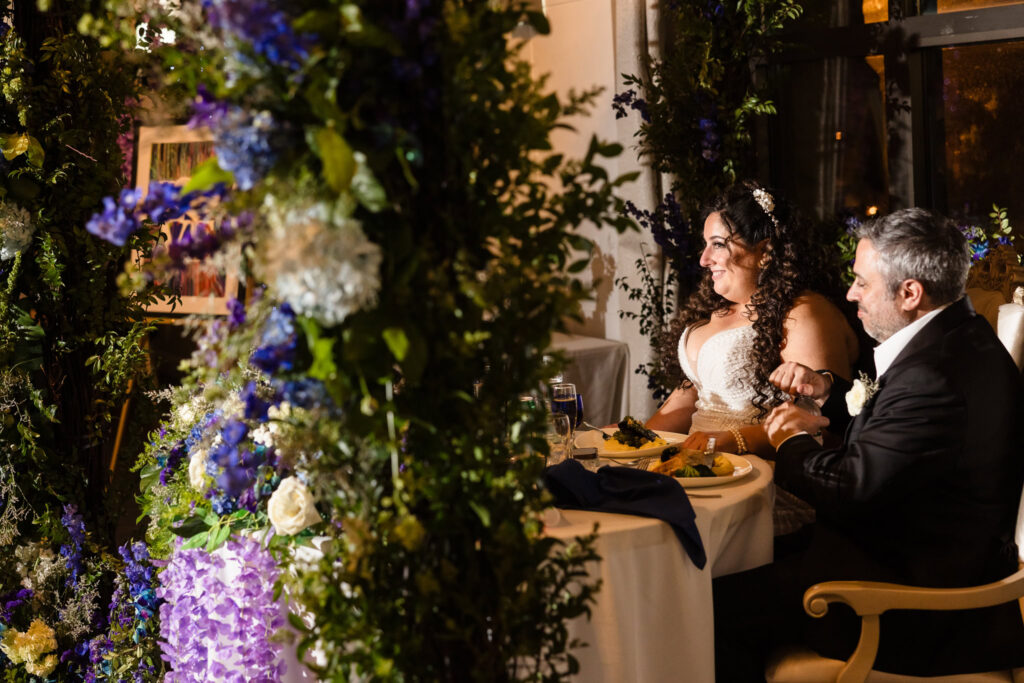 Bride and groom seated at their sweetheart table, surrounded by floral designs and candles, sharing a quiet laugh.
