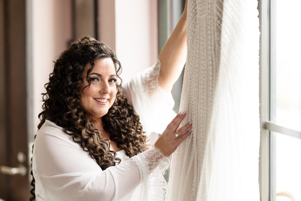 Bride smiling as she gently touches her gown near a window, lit by natural light before the ceremony.