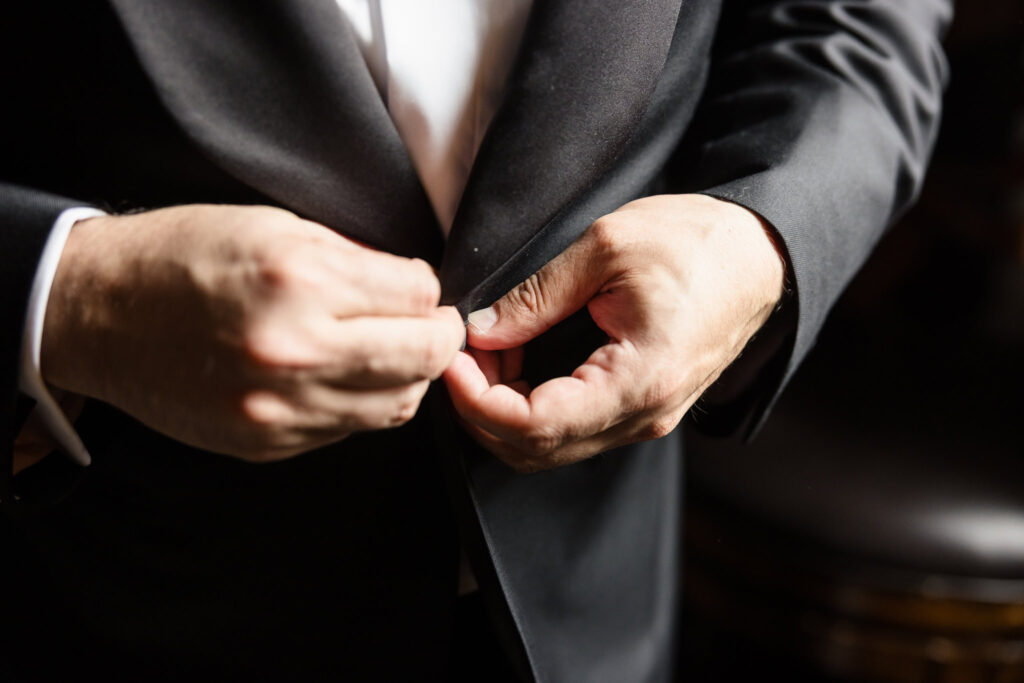 Groom buttoning his tuxedo jacket with a sense of calm and focus in a warmly lit room.