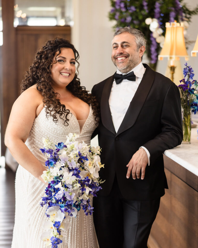 Bride and groom smiling together inside the venue, with the bride holding a white and purple bouquet.