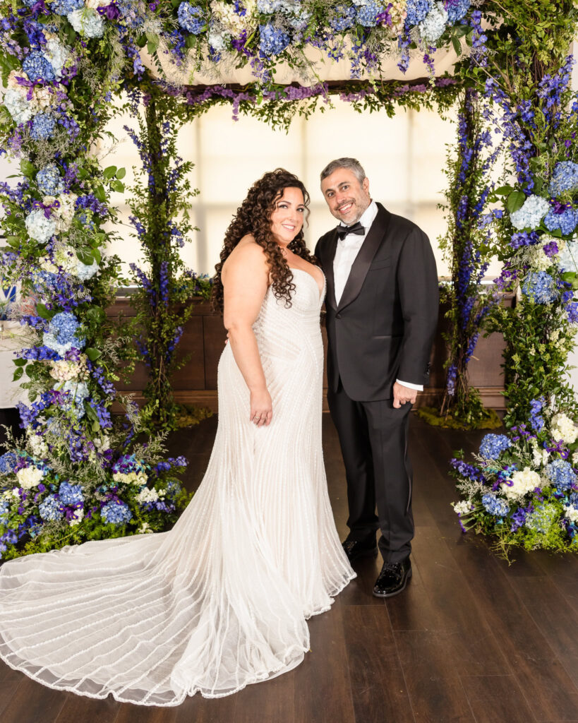 Bride and groom standing beneath a floral arch bursting with purple and white flowers, just before the ceremony.