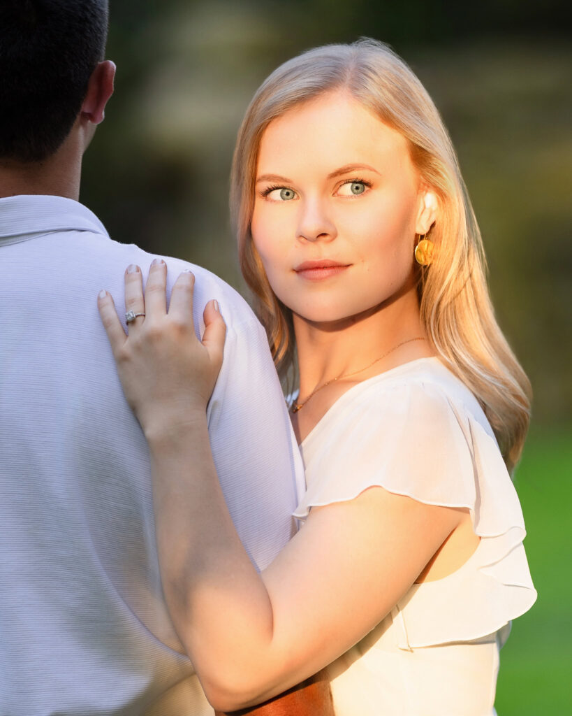 Bride-to-be looking toward the camera over her fiancé’s shoulder during golden hour at Greenwich Point Park.