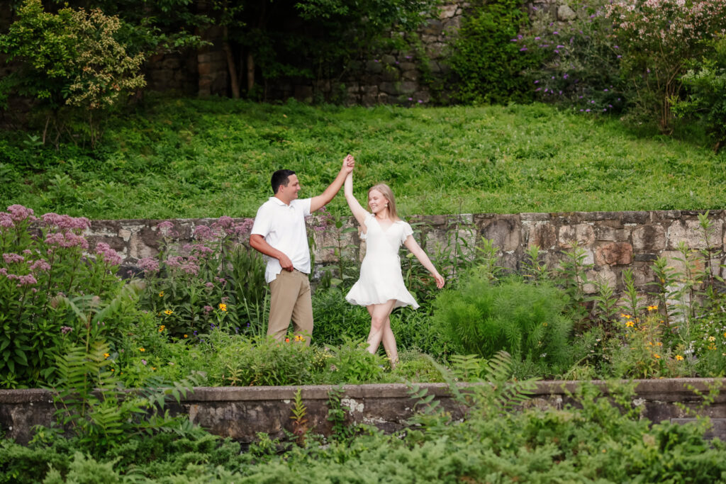 Couple twirling in lush summer greenery along a stone terrace at Greenwich Point Park; her white dress swirls as he lifts her hand.